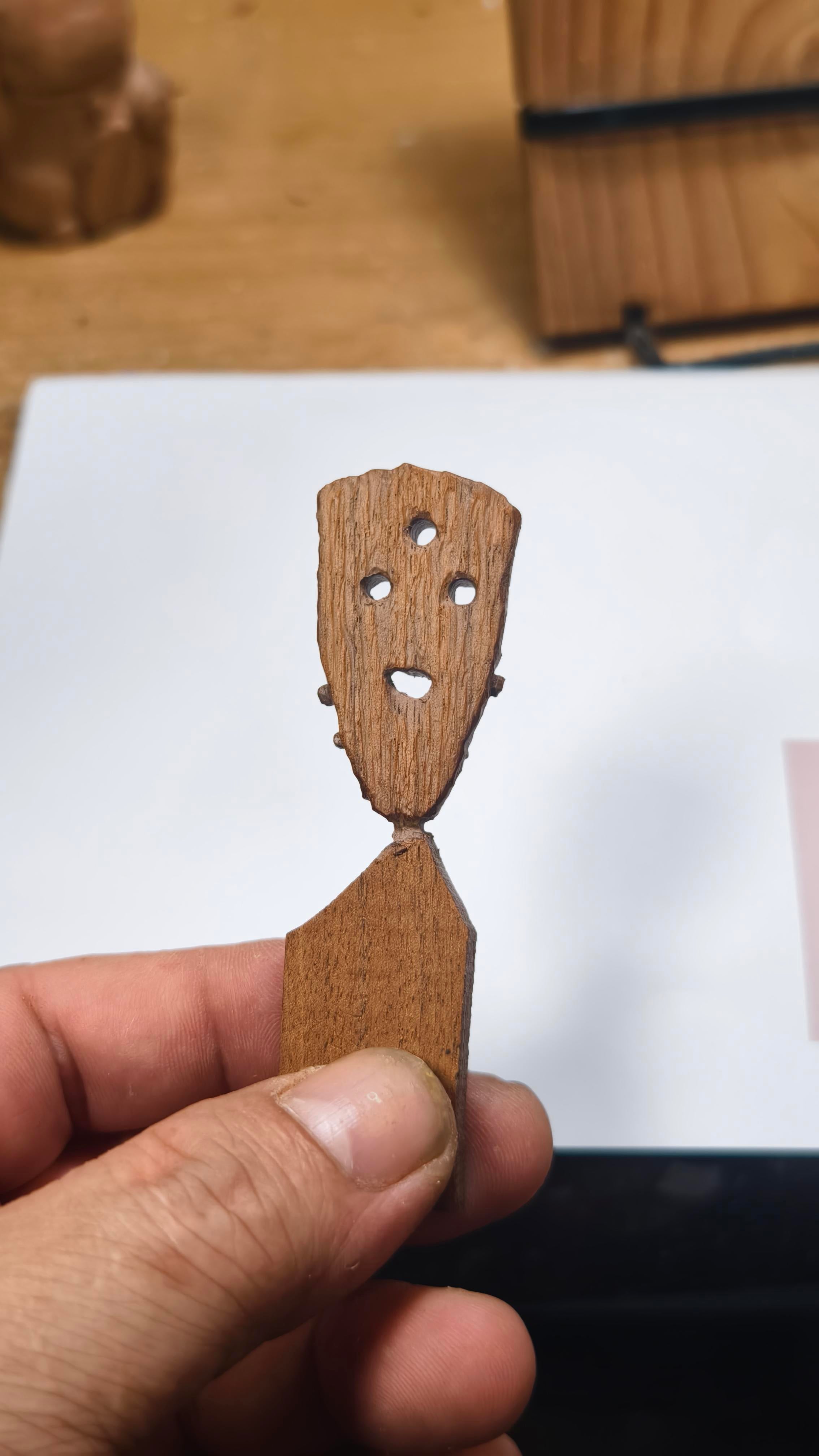 Wooden cutout of a face with holes for eyes, nose, and mouth held by a hand on a light-colored surface. Gríma