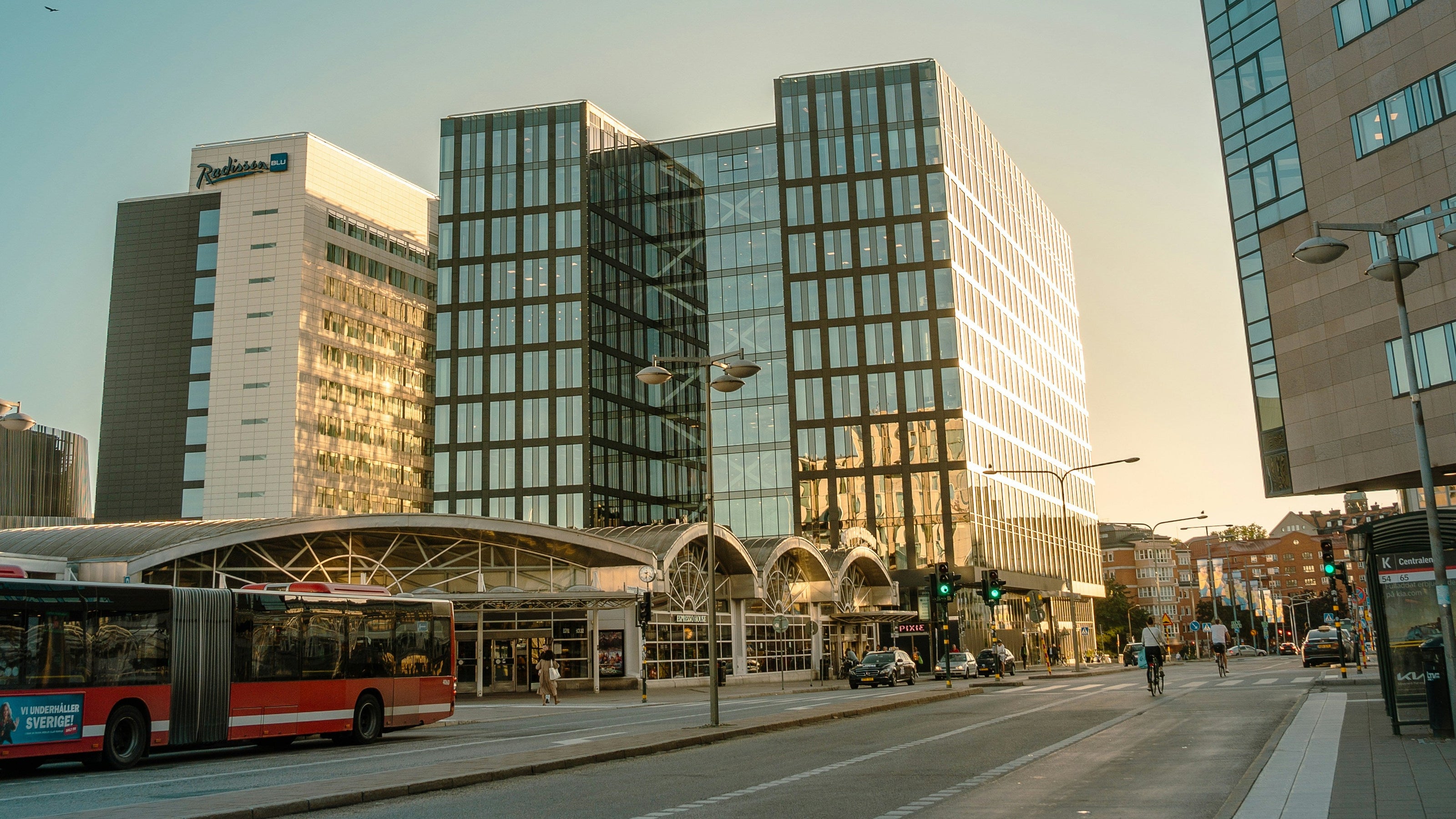 A wide-angle street scene in a city at sunset, featuring modern glass office buildings, a bus stop, a red and white bus on the left, and a sidewalk on the right under a clear blue sky.