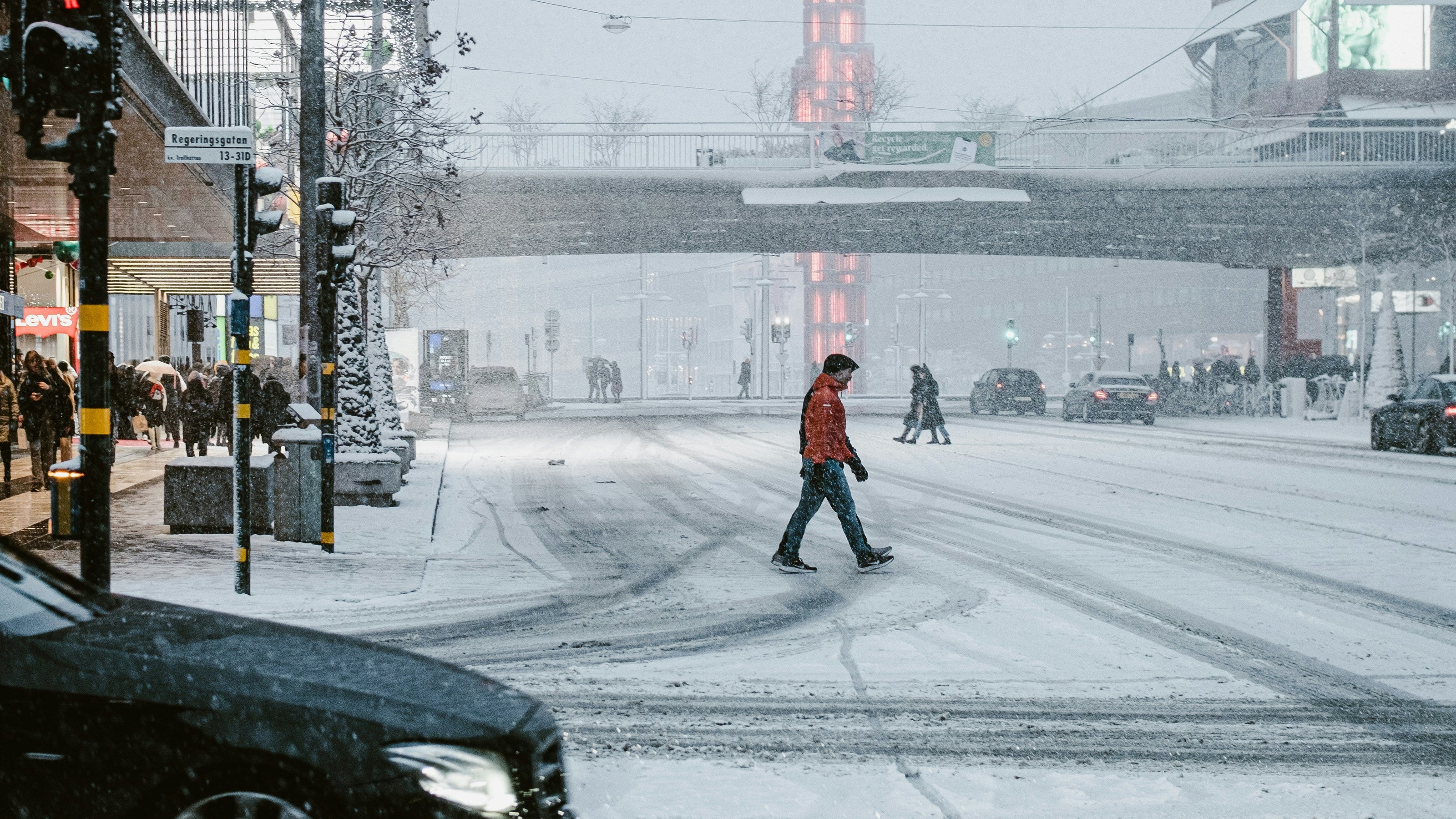 A snowy urban street scene with a pedestrian crossing a snow-covered crosswalk in the distance, framed by buildings on both sides and a red-lit bridge or tower in the background. A black car is partially visible in the lower left foreground.