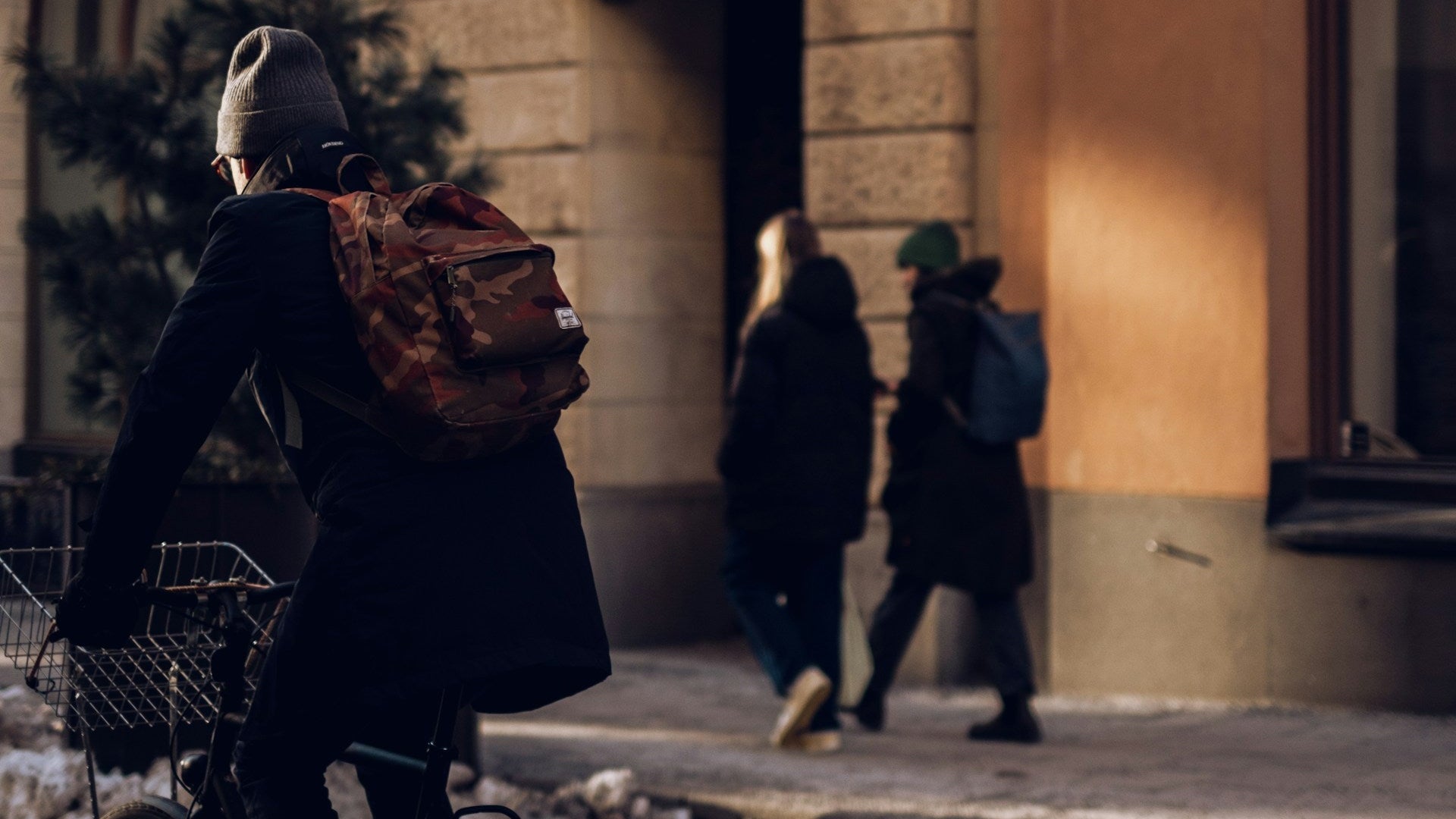 A person riding a bicycle in a city street in front of a stone building, wearing a dark coat, gray beanie, and a brown backpack. Snow is piled along the curb, and two pedestrians walk on the sidewalk in the background.