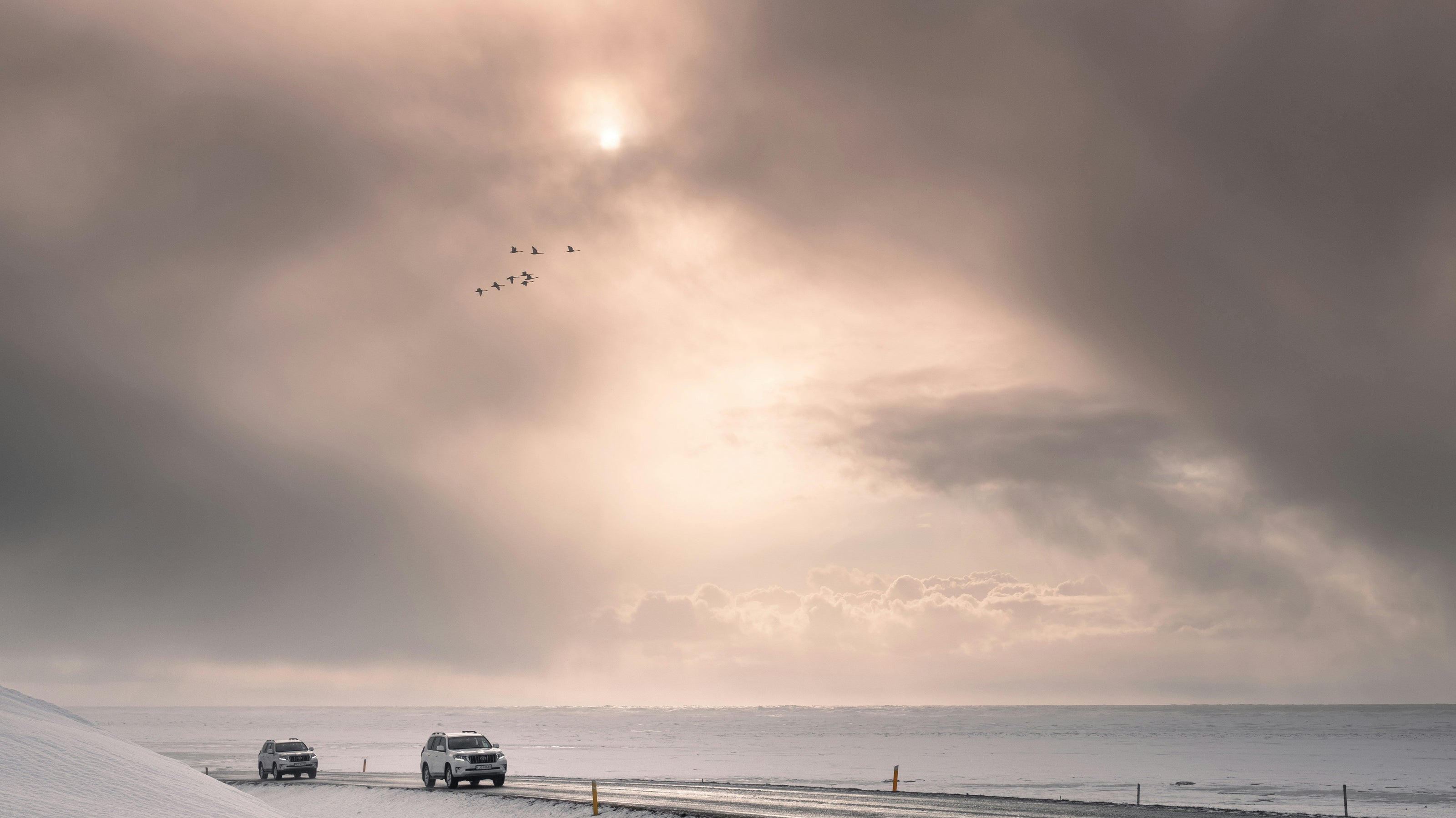 A dramatic snowy landscape with two cars driving along a road beside a frozen body of water under a cloudy sky with the sun shining through. A small flock of birds is visible in the sky.