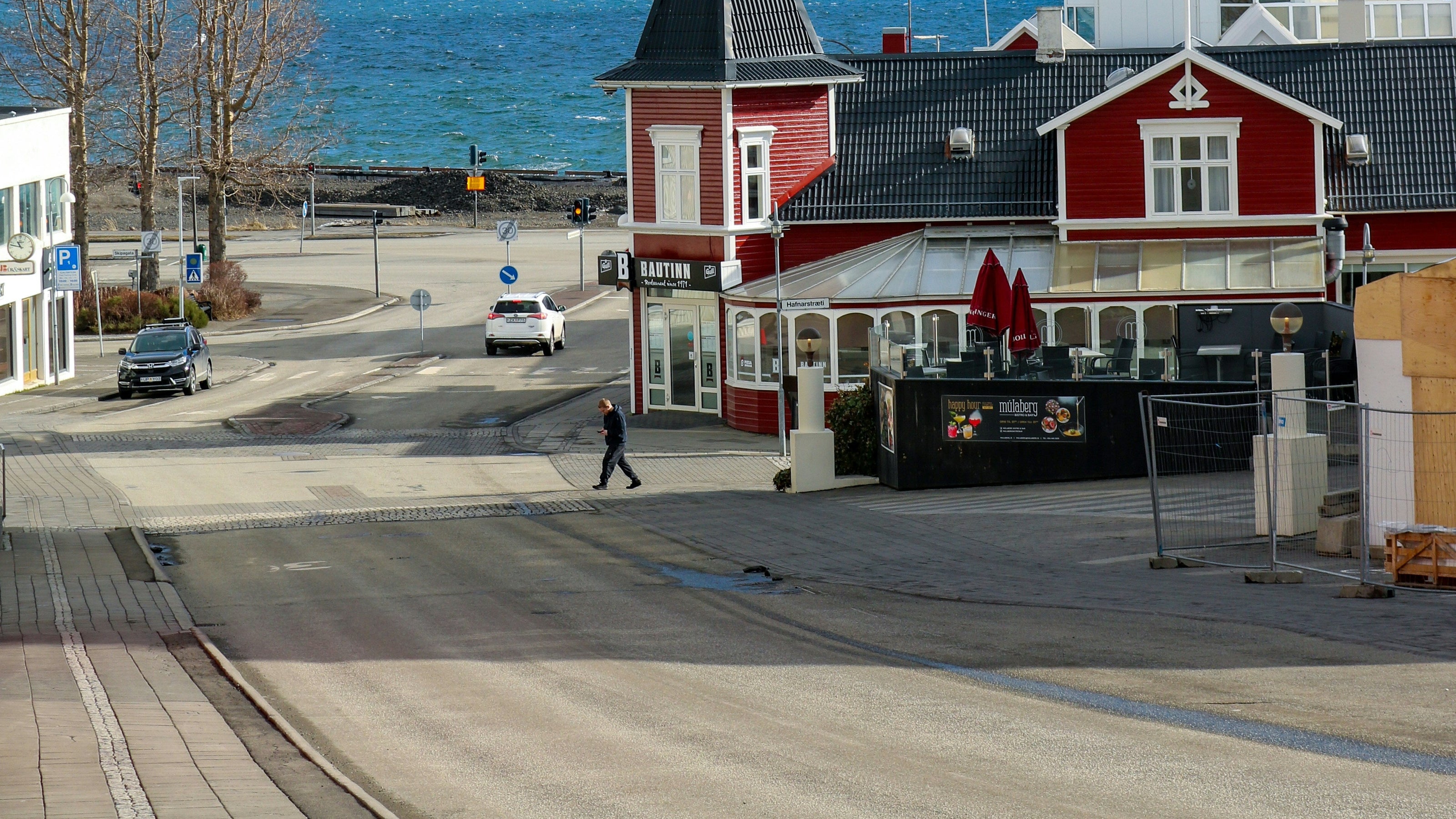 A scenic view of a coastal town with red and white buildings in the foreground, a blue body of water, and snow-covered mountains under a clear blue sky.