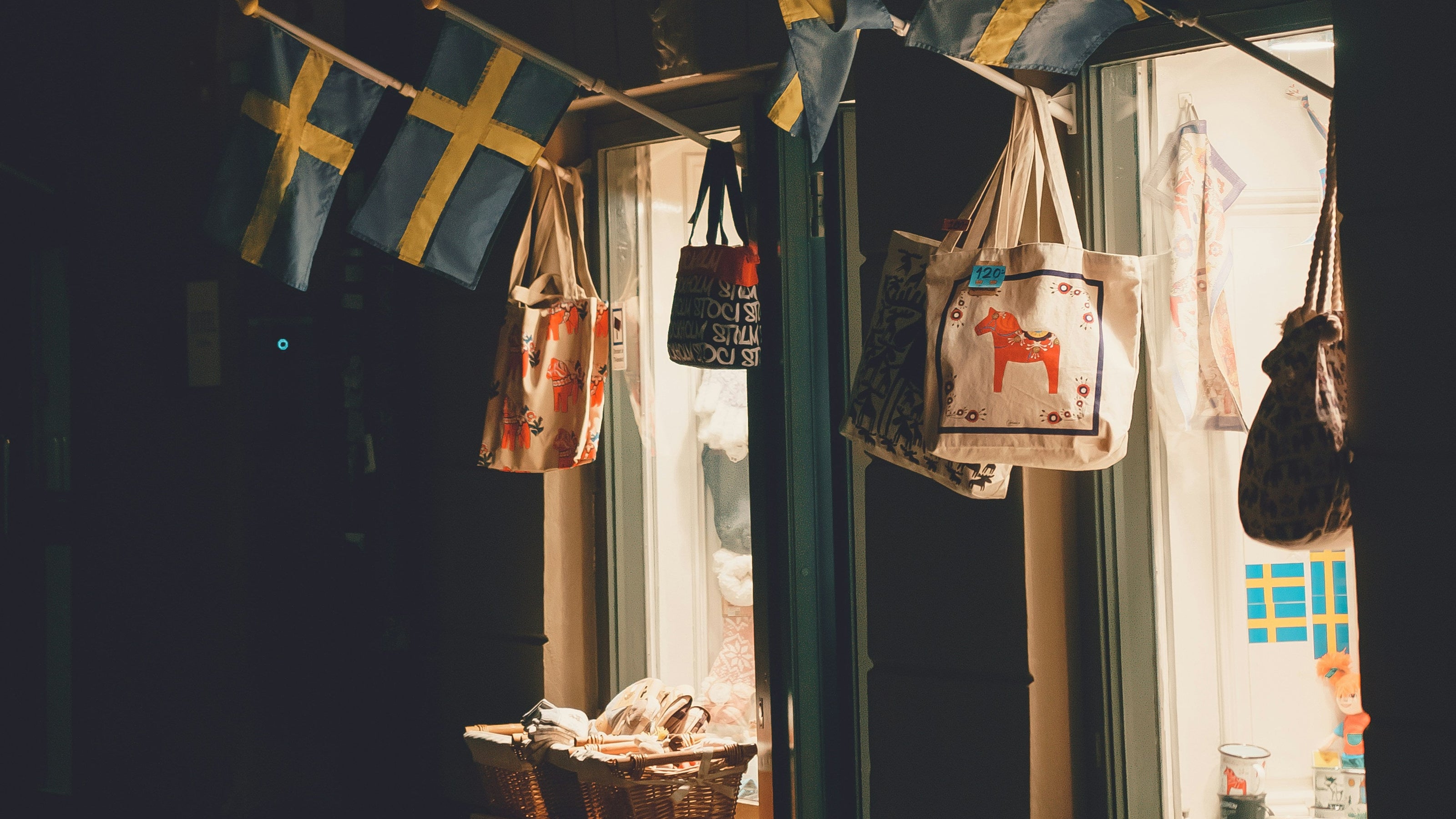 A storefront window at night with Swedish flags hanging from a pole, tote bags, and baskets displayed outside. A sign above reads 'OLD TOWN GIFTS'.