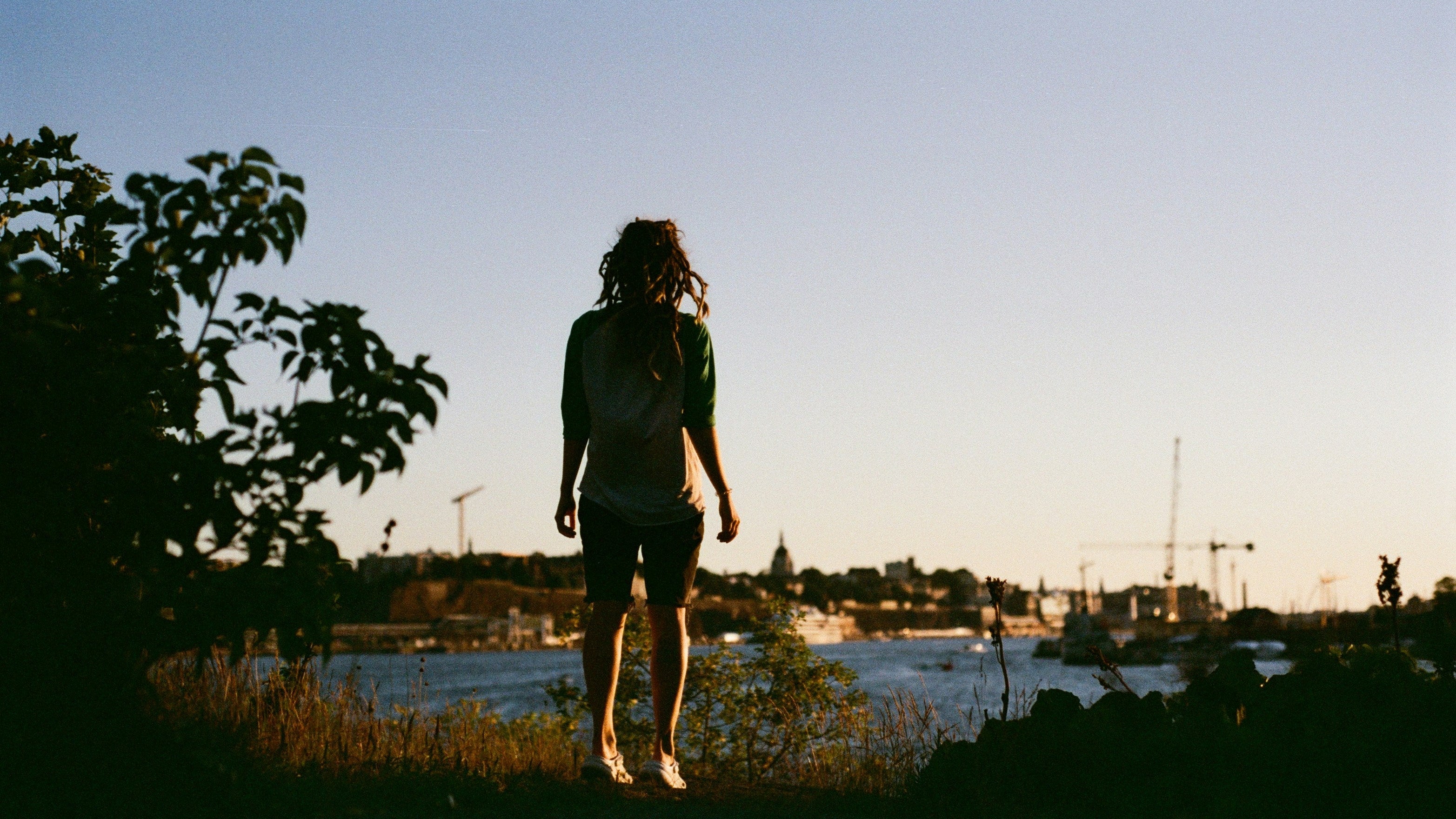 A person standing outdoors by a body of water at sunset, seen from behind, with trees in the foreground and a distant shoreline with cranes and buildings.